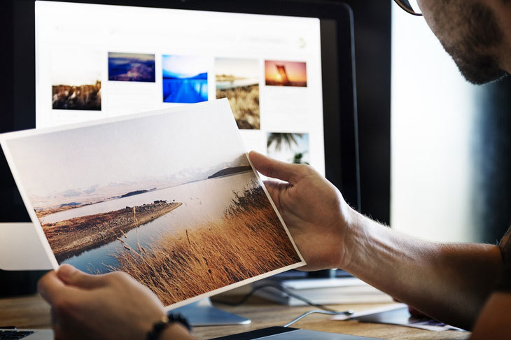 Man wearing an eyeglasses is holding a printed photo and in front of his is a computer desktop where he edit all photos using the adobe photoshop elements 15 